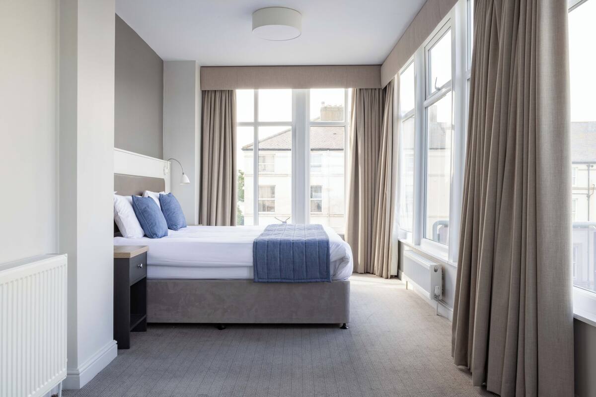 A modern hotel room with a large bed, a bedside table, a lamp, and a view of the city through large windows.