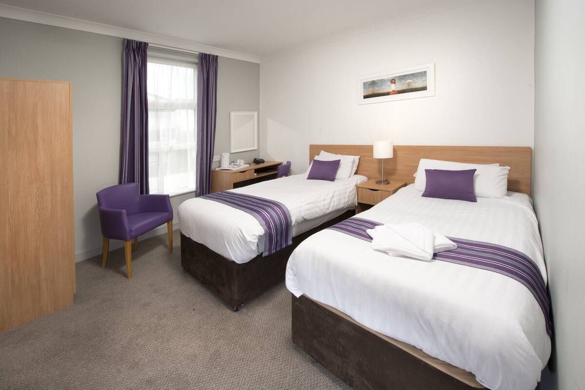 A hotel room with two beds, a desk, a chair, and a window with curtains.