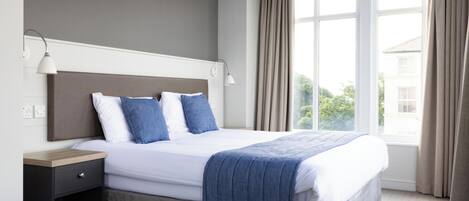Una cama bien hecha con una manta azul, una mesita de noche con una lámpara y una ventana con cortinas.
