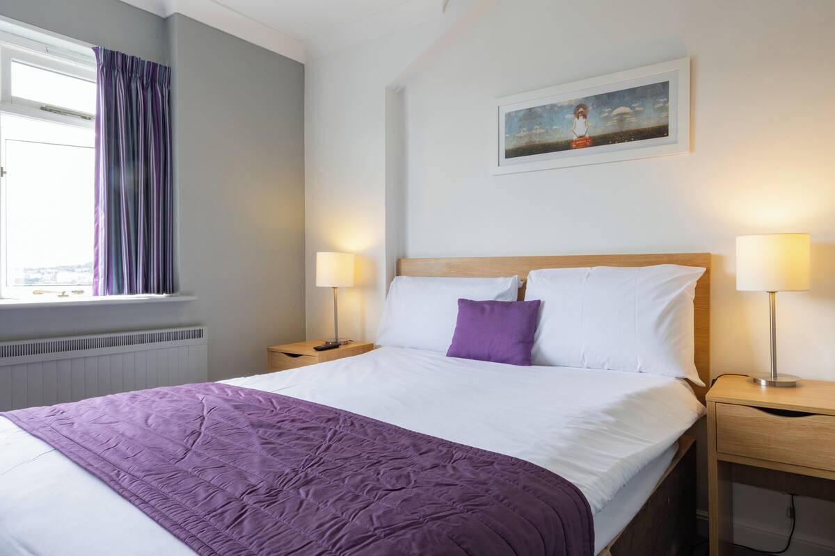 A neatly made bed with a purple blanket, two bedside lamps, and a painting on the wall.