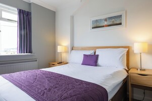 Una cama bien hecha con un edredón morado, dos lámparas de noche y un cuadro en la pared.