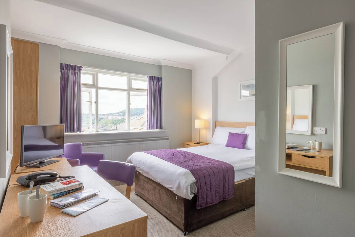 A hotel room with a bed, desk, chair, and a large window with curtains.