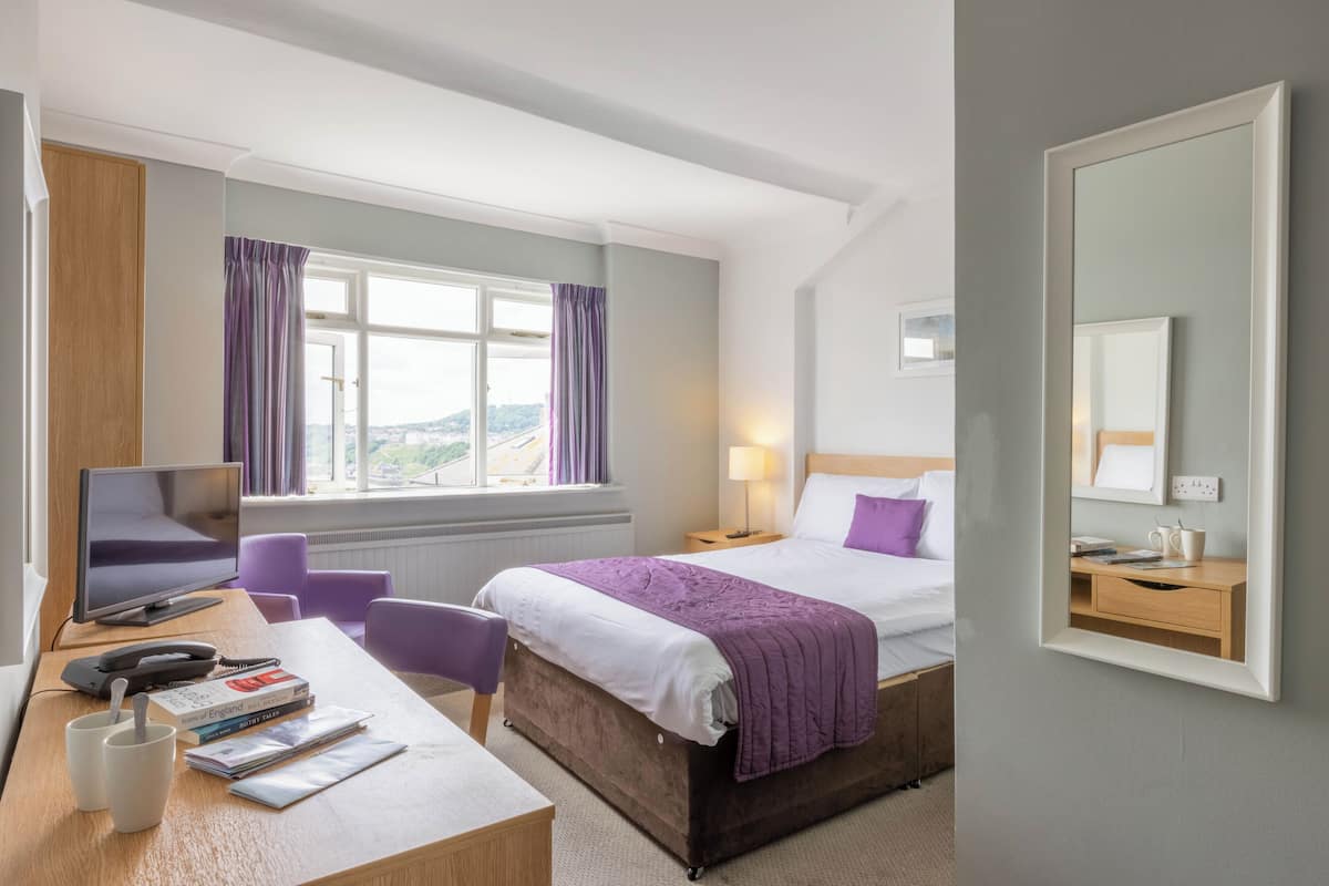 A hotel room with a bed, desk, chair, and a large window with curtains.