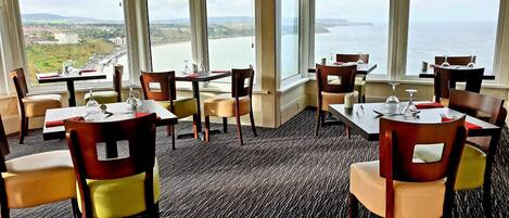 A dining area with tables and chairs, large windows with a view of the sea and a distant landscape.