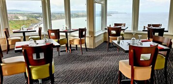 Un comedor con mesas y sillas, ventanas amplias con vista al mar y un paisaje distante.