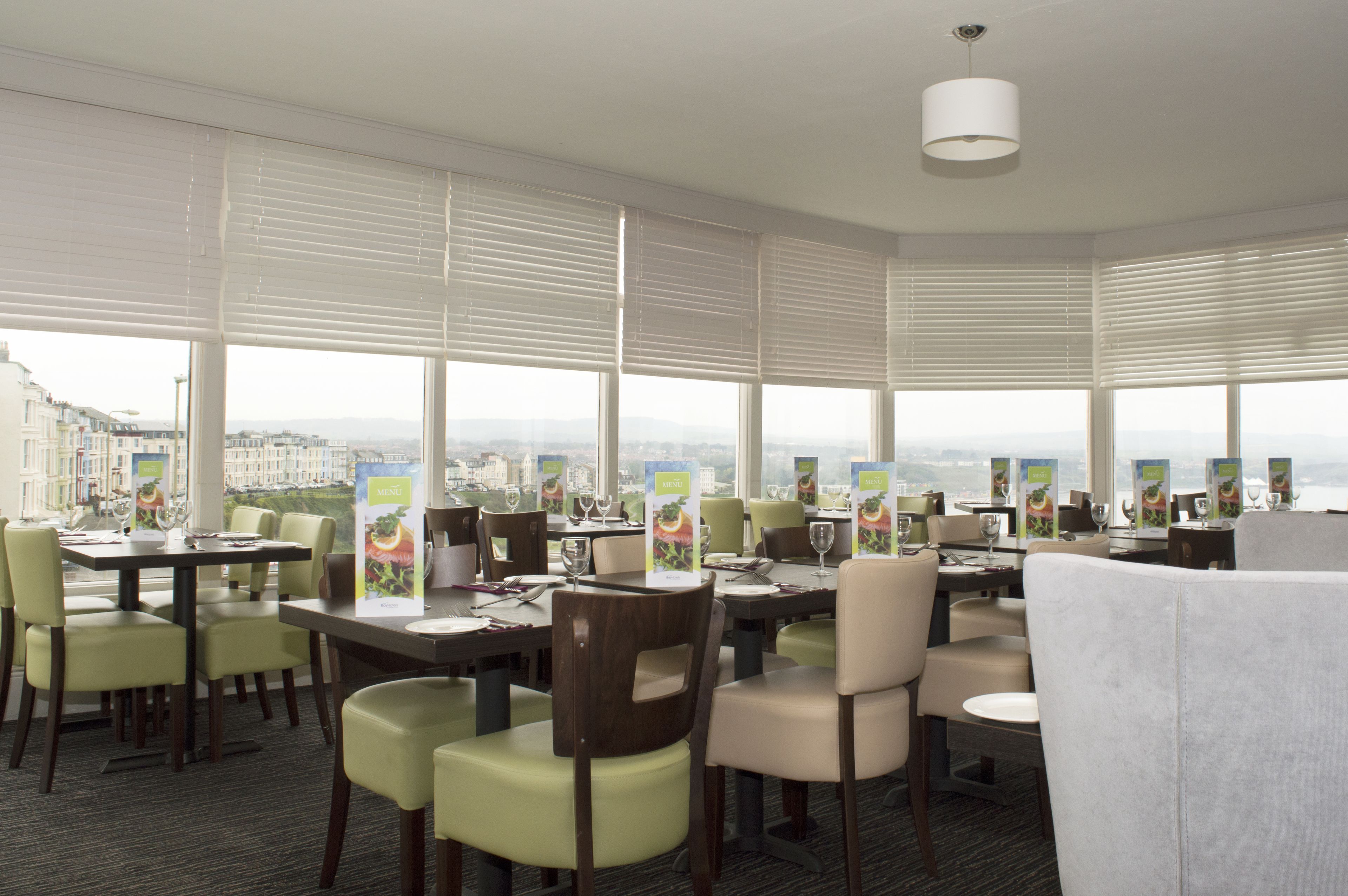 A restaurant with multiple tables and chairs, large windows with blinds, and a view of a cityscape.