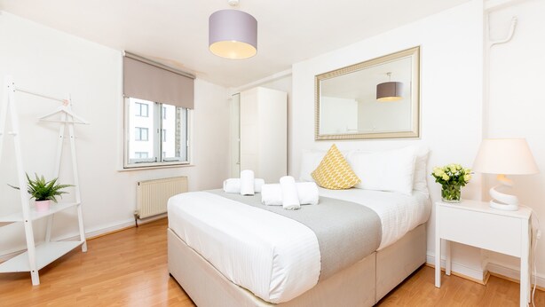 A modern bedroom with a large bed, a bedside table with a lamp, a mirror, and a window with a view of buildings. - Heart of Shoreditch Apartments  (London)