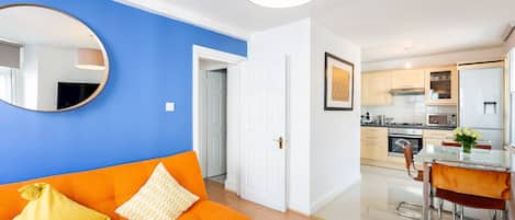 Un apartamento moderno con pared azul de acento, un espejo redondo, un sofá naranja, un comedor con mesa de vidrio y una cocina con electrodomésticos de acero inoxidable.
