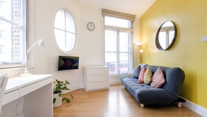 A modern living room with a white desk, a blue sofa with colorful cushions, a round mirror, and a television on the wall. - Heart of Shoreditch Apartments  (London)