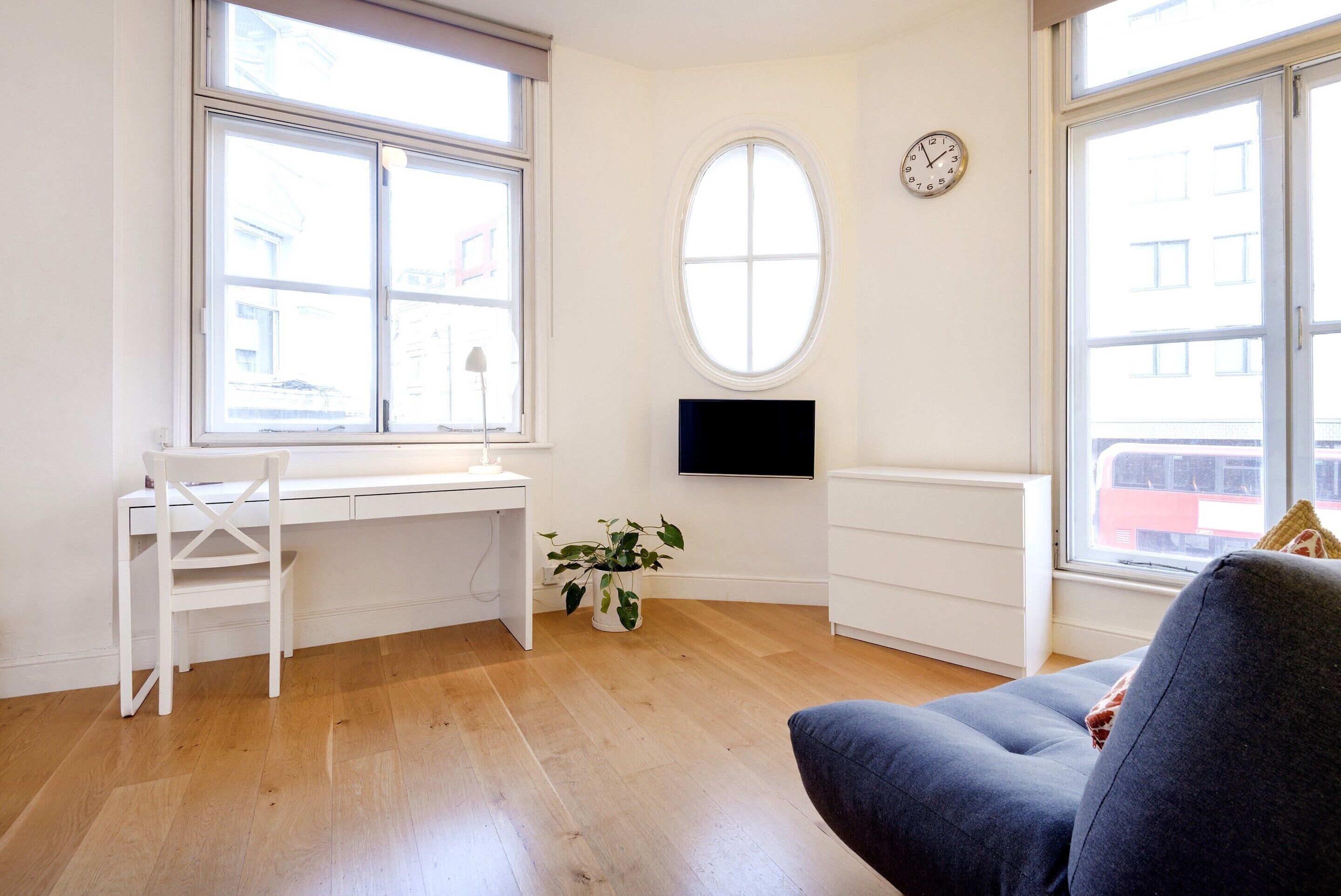Una habitación con un escritorio blanco, una silla, una planta en maceta, un televisor en la pared y ventanas amplias.
