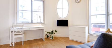 Una habitación con un escritorio blanco, una silla, una planta en maceta, un televisor en la pared y ventanas amplias.
