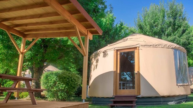 A single-story yurt with a wooden door and a covered entrance, situated on a grassy area with a wooden deck and picnic table. - Camping le Beauvillage - Yourte (Lauzerte)