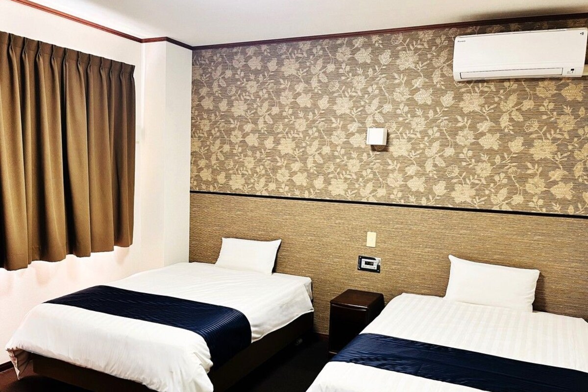 A hotel room with two beds, a bedside table, an air conditioning unit, and a window with curtains.