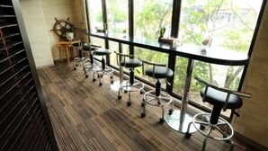A row of bar stools with wheels, a ship's wheel decoration, and a view of greenery through large windows.