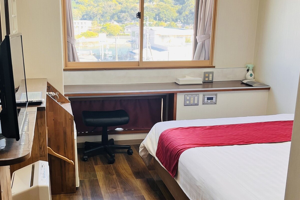 A hotel room with a bed, desk, chair, and window with a view of greenery.