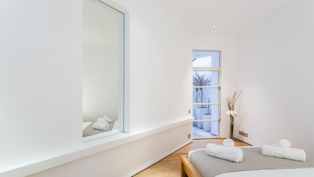 A modern bedroom with a large window, a bed with white linens, and a small bedside table with a vase of flowers.