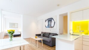 A modern living room with a sofa, coffee table, and a vase of flowers. There is a kitchen area with a yellow-lit counter and cabinets.