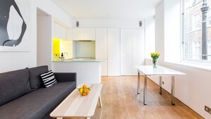 A modern apartment with a grey sofa, a wooden coffee table with fruit, a white dining table, and a kitchen in the background.