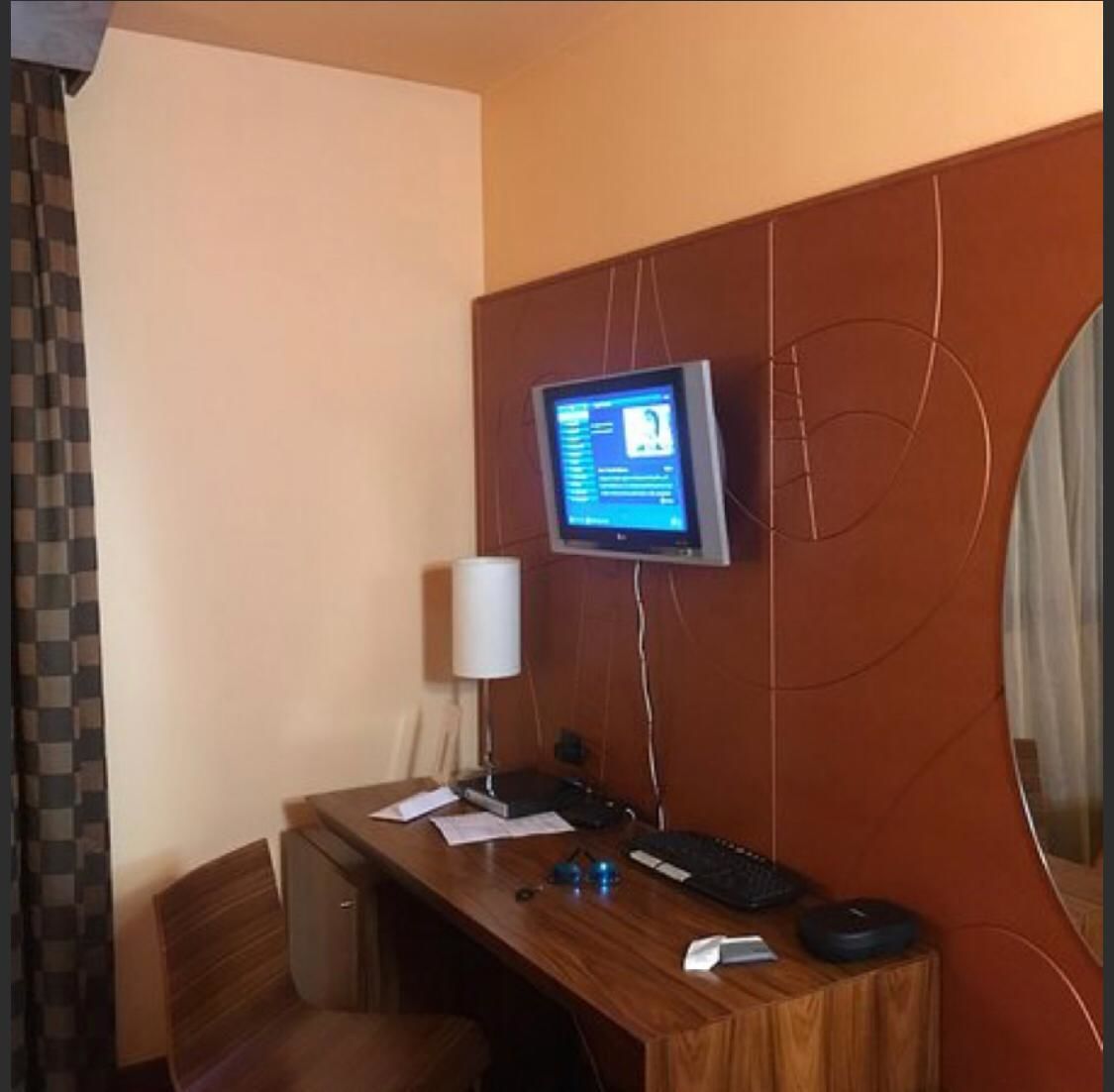 A hotel room with a wooden desk, a computer monitor, a lamp, and a chair.