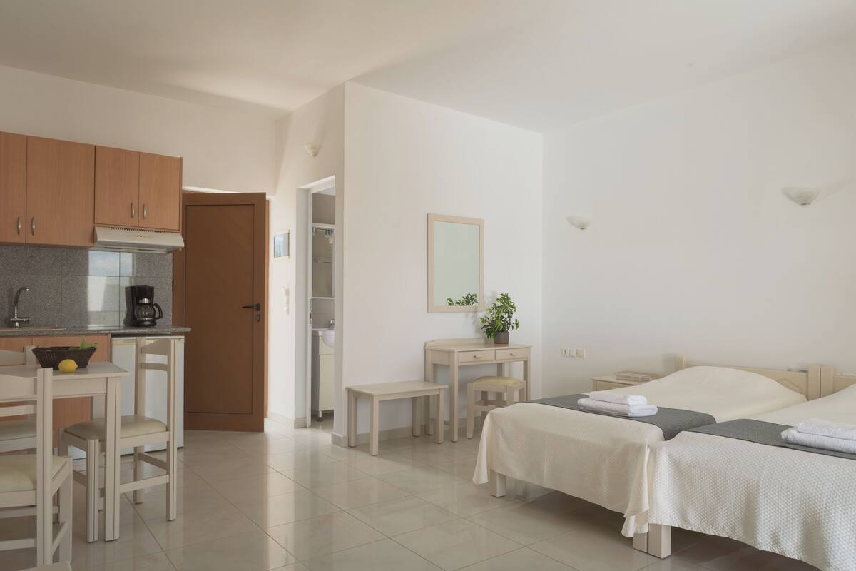 A hotel room with a kitchenette, a bed, a bedside table, and a small desk with a chair.