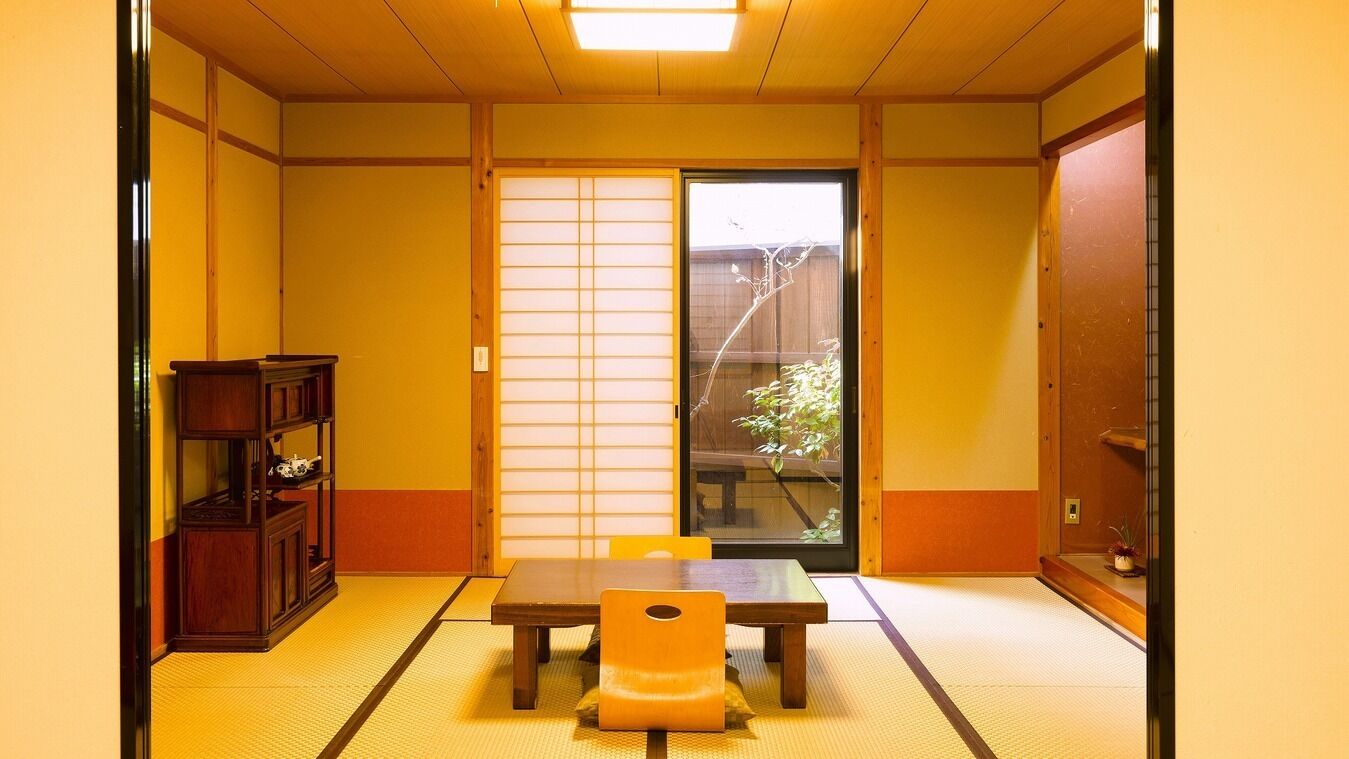 Ein traditioneller japanischer Raum mit Tatami-Boden, einer Schiebetür, einem Holztisch und einem Regal mit Dekoration.