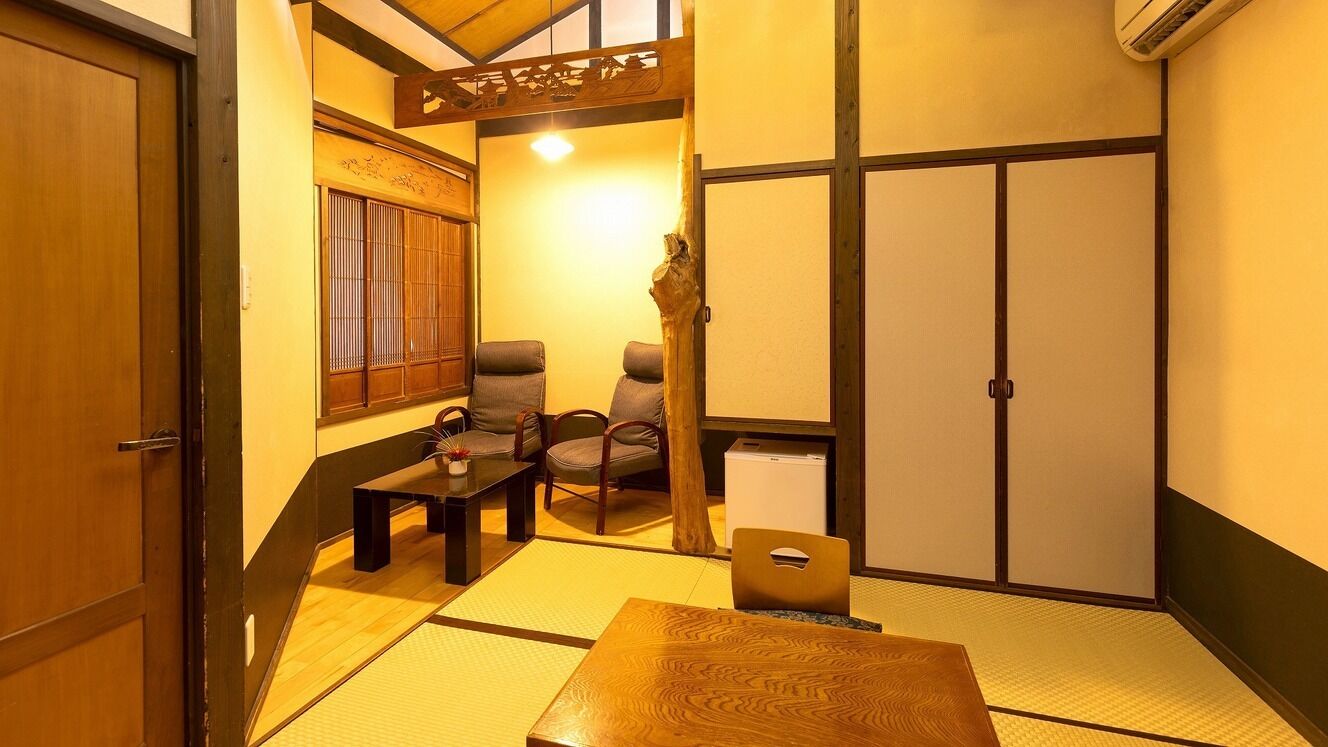 Ein traditioneller japanischer Raum mit Tatami-Boden, einem niedrigen Tisch, zwei Stühlen, einem kleinen Tisch mit einer Vase und einer Schiebetür.