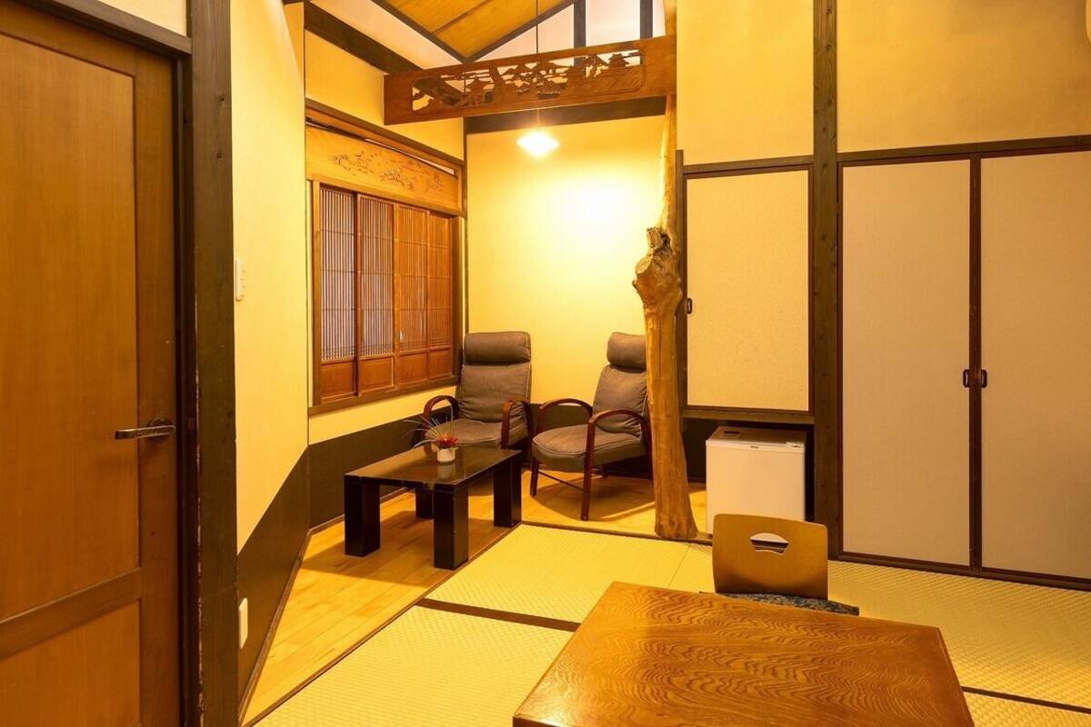 A traditional Japanese room with tatami flooring, a low table, two chairs, a small table with a vase, and a sliding door.