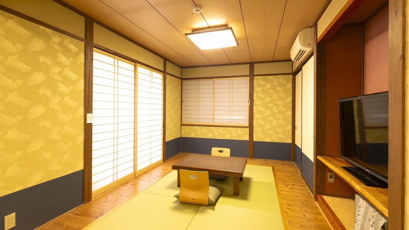 Ein traditioneller japanischer Raum mit Tatami-Böden, einem niedrigen Tisch, einem Stuhl, Schiebetüren aus Shoji-Papier und einem Fernseher.