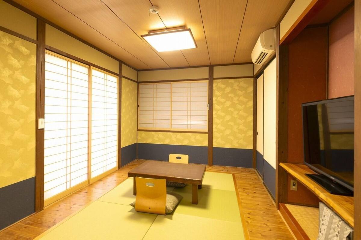 A traditional Japanese room with tatami flooring, a low table, a chair, sliding shoji doors, and a television.