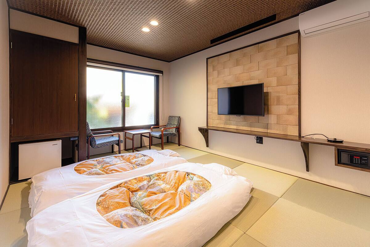 A hotel room with tatami flooring, a bed with a patterned bedspread, a television mounted on a brick wall, and a chair by the window.