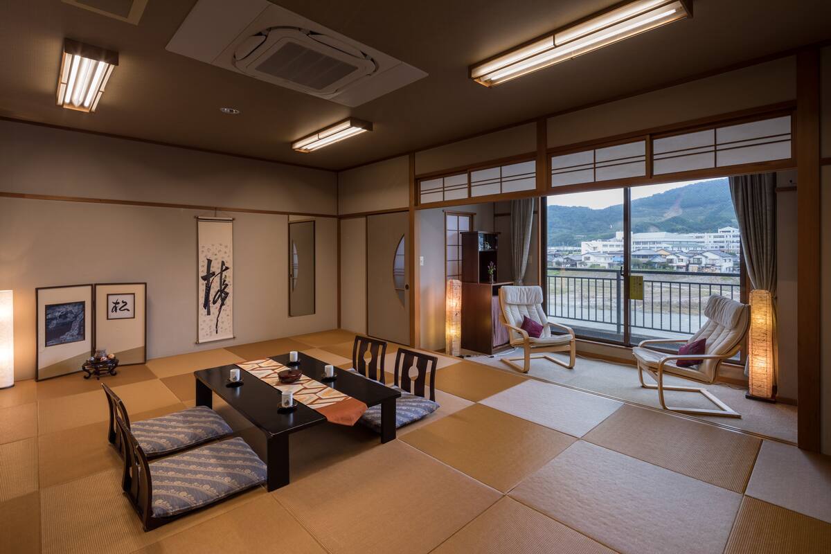 A traditional Japanese room with tatami flooring, a low table with cushions, and a view of a cityscape through large windows.