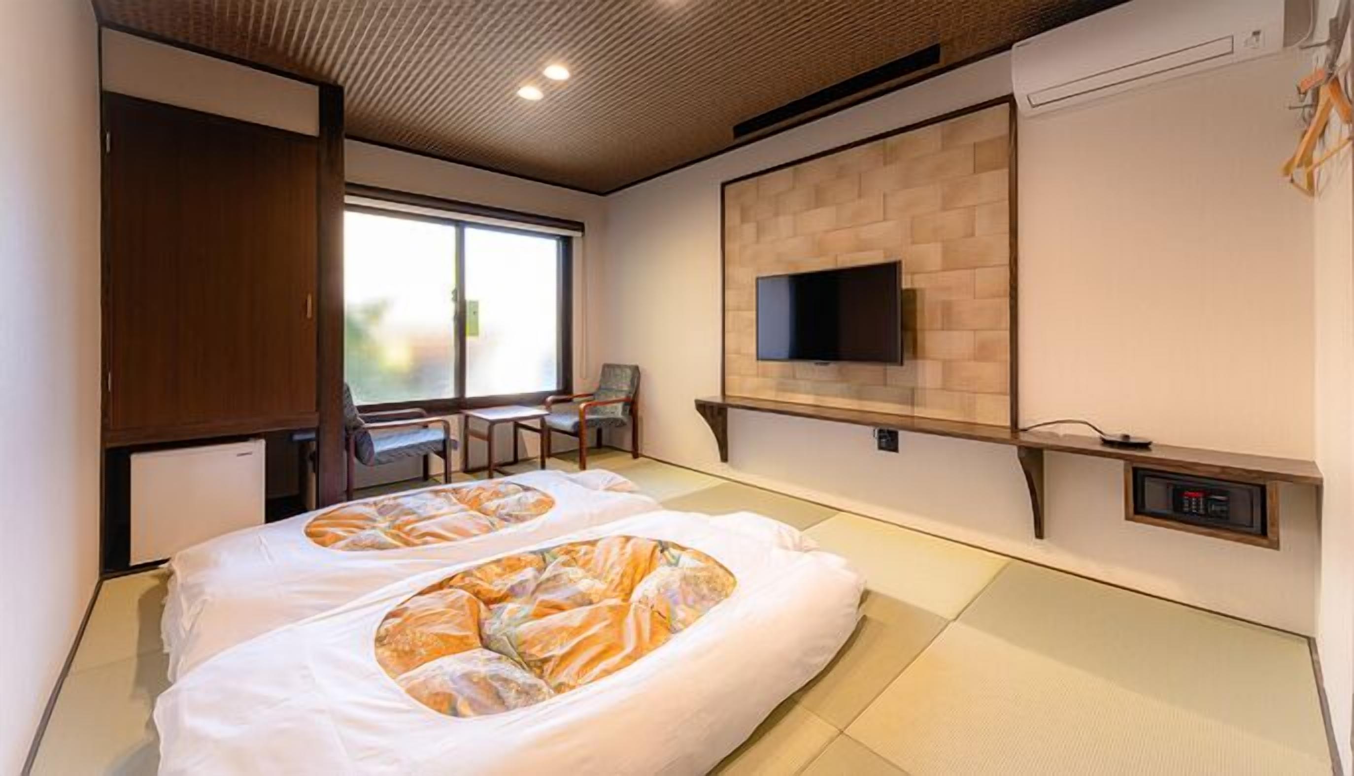 A hotel room with tatami mats, a television mounted on a brick wall, and a desk with chairs.