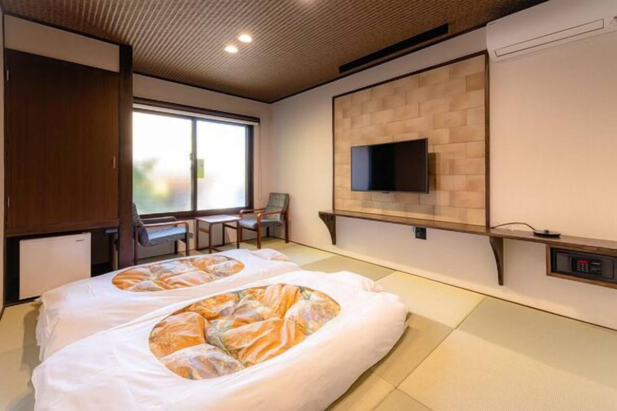 A hotel room with tatami mats, a television mounted on a brick wall, and a desk with chairs.