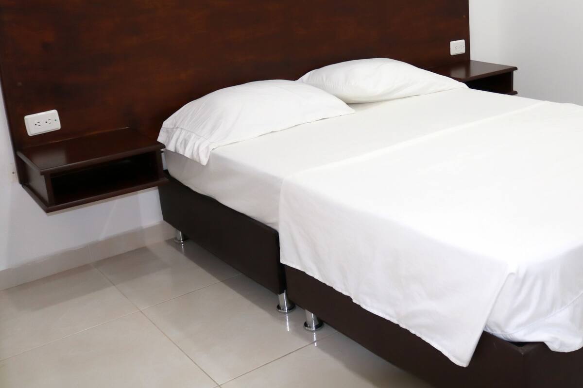 A neatly made bed with white linens, a wooden headboard, and a small bedside table with a power outlet.