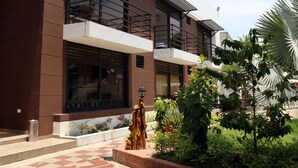 A modern building with a brown facade, large windows, and a small garden area with plants and a decorative tree sculpture. - Hotel Restaurante Versalles (Melgar)