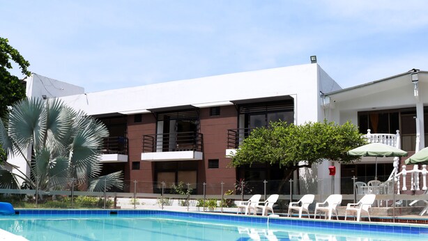 A swimming pool with lounge chairs and umbrellas, surrounded by buildings with balconies and a tree. - Hotel Restaurante Versalles (Melgar)