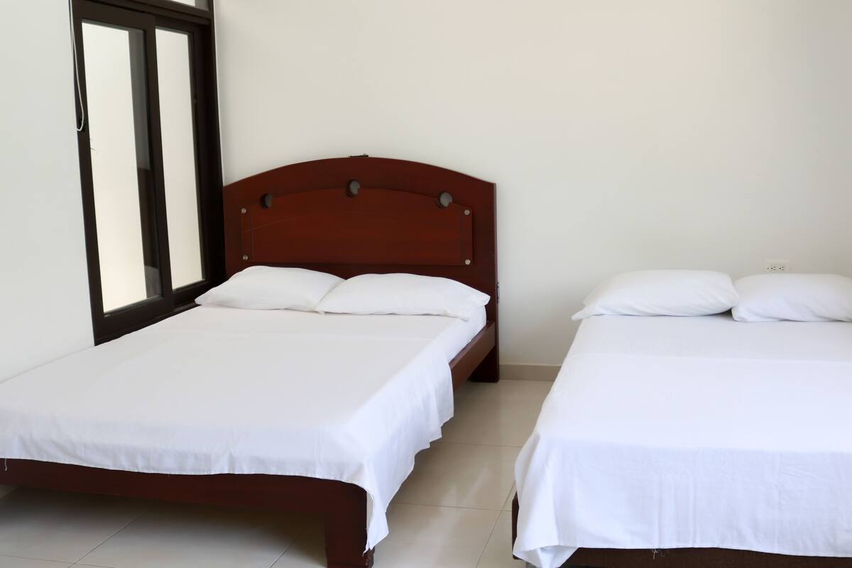 A room with two beds, a window with blinds, and an air conditioning unit on the wall.