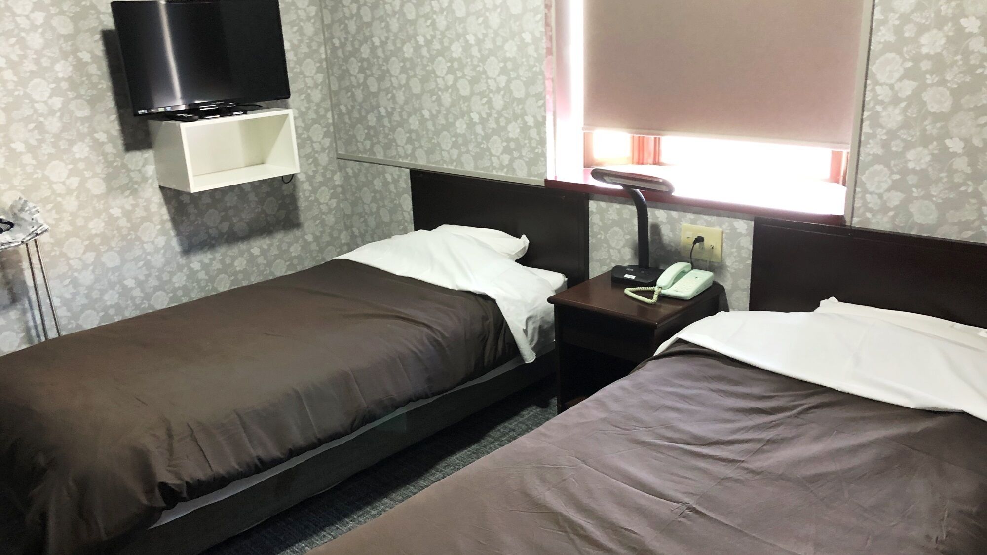 Ein Hotelzimmer mit zwei Einzelbetten, einem Nachttisch mit Telefon, einem an einer Ablage montierten Fernseher und einem Fenster mit Vorhängen.