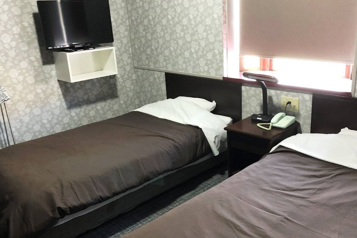 A hotel room with two single beds, a bedside table with a phone, a TV mounted above a shelf, and a window with curtains.