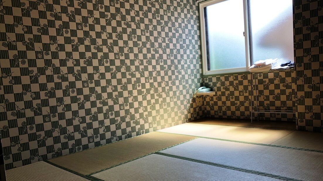 Ein Raum mit Tatami-Matten, einem karierten Wandmuster, einem Fenster und einem kleinen Regal mit Gegenständen.
