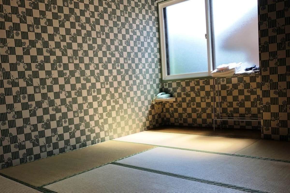 Una habitación con suelo de tatami, pared con diseño de cuadros, una ventana y una pequeña estantería con objetos.