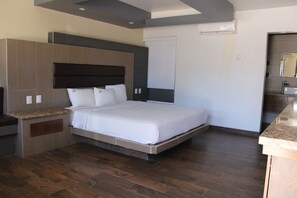 A modern hotel room with a large bed, built-in headboard, and a small kitchenette area.