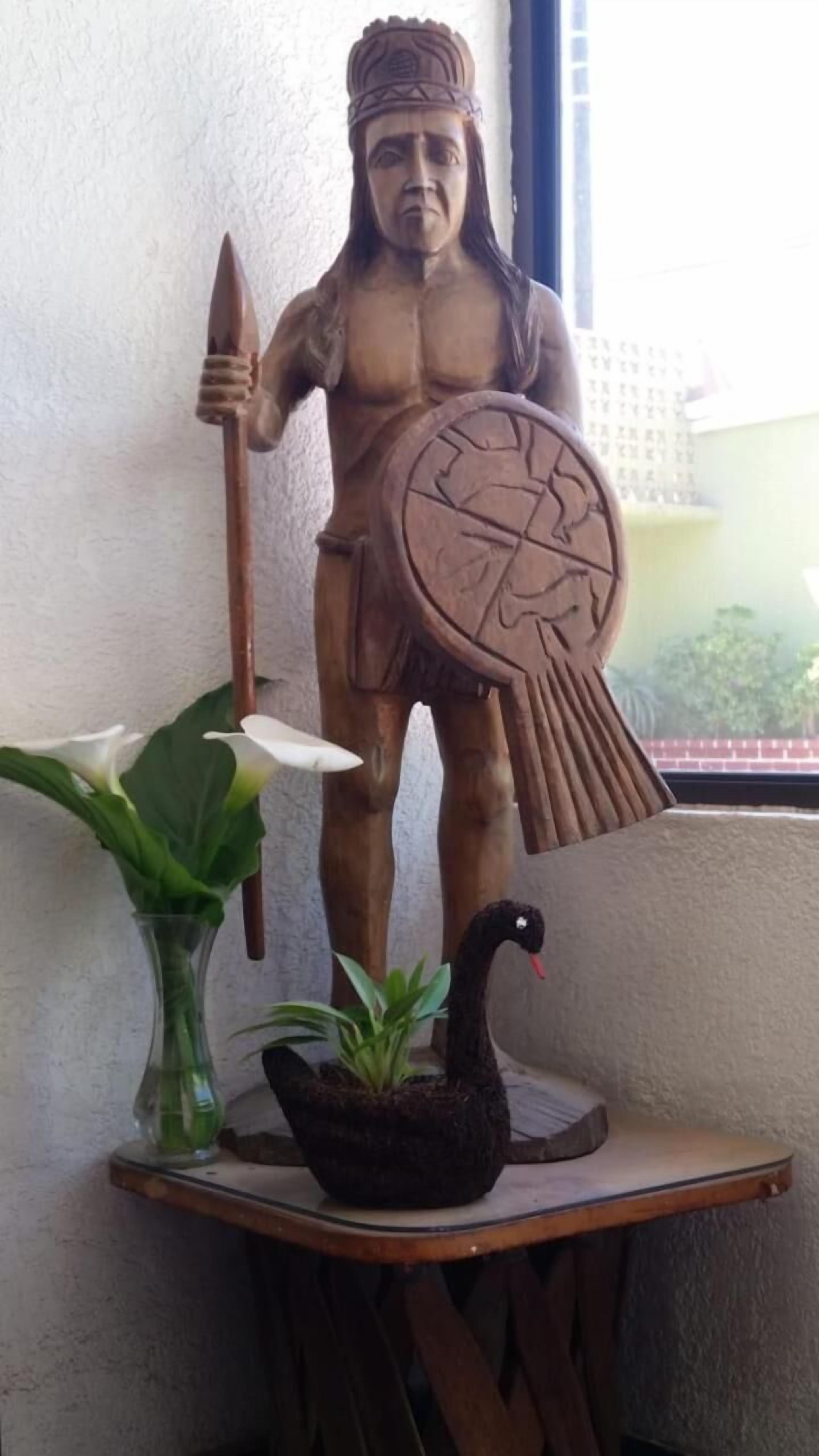 A statue of a warrior holding a spear and shield, placed on a wooden table with a black swan and a white flower beside it.