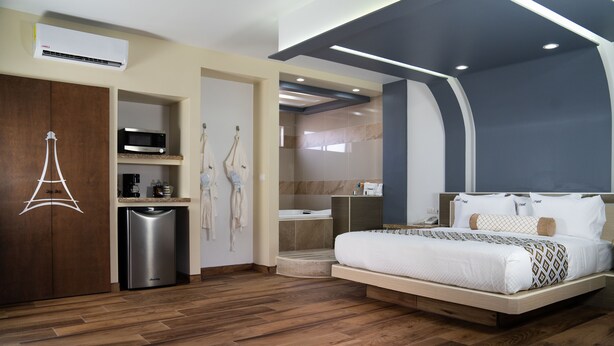 A modern hotel room with a wooden floor, a bed with white bedding, a minibar, a microwave, and a shower area. - Hotel Eiffel (Ensenada)
