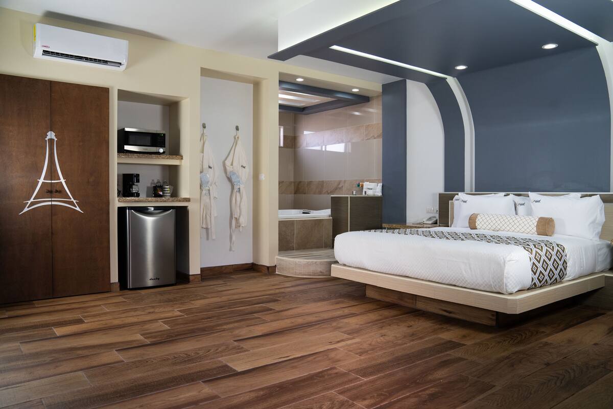 Habitación de hotel moderna con piso de madera, cama con ropa blanca, minibar, microondas y zona de ducha.