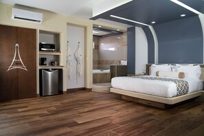 Habitación de hotel moderna con piso de madera, cama con ropa blanca, minibar, microondas y zona de ducha.
