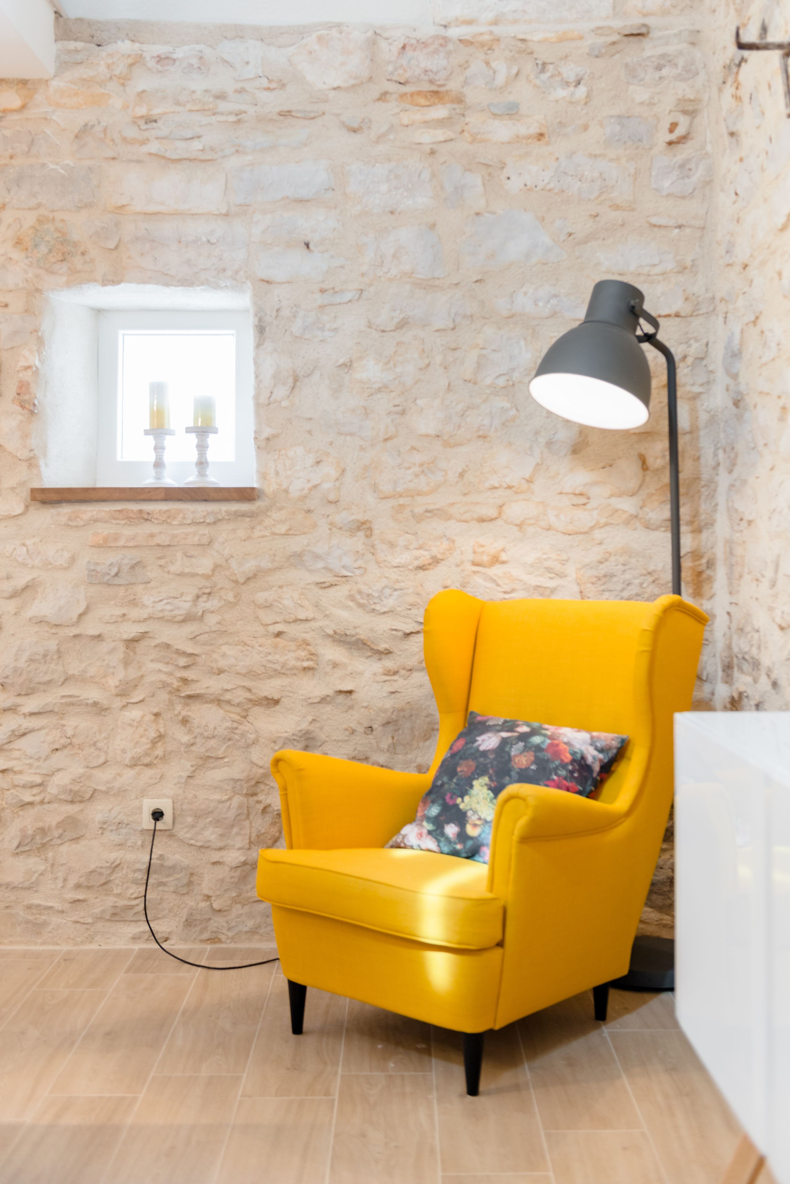 A room with a stone wall, a yellow armchair with a floral pillow, a black floor lamp, and a small window with candles.