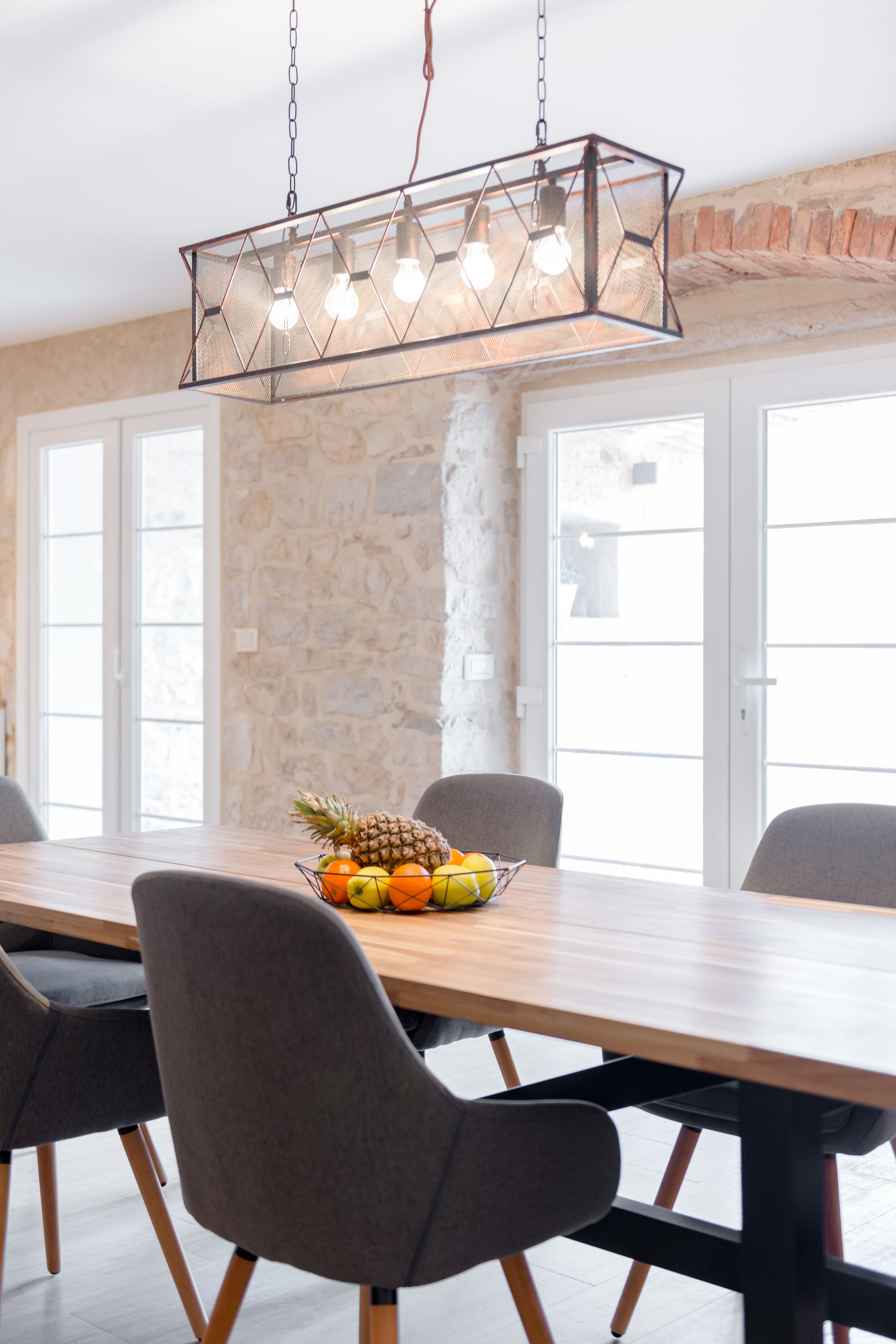 A dining area with a long wooden table, six chairs, a fruit bowl, and a hanging light fixture.