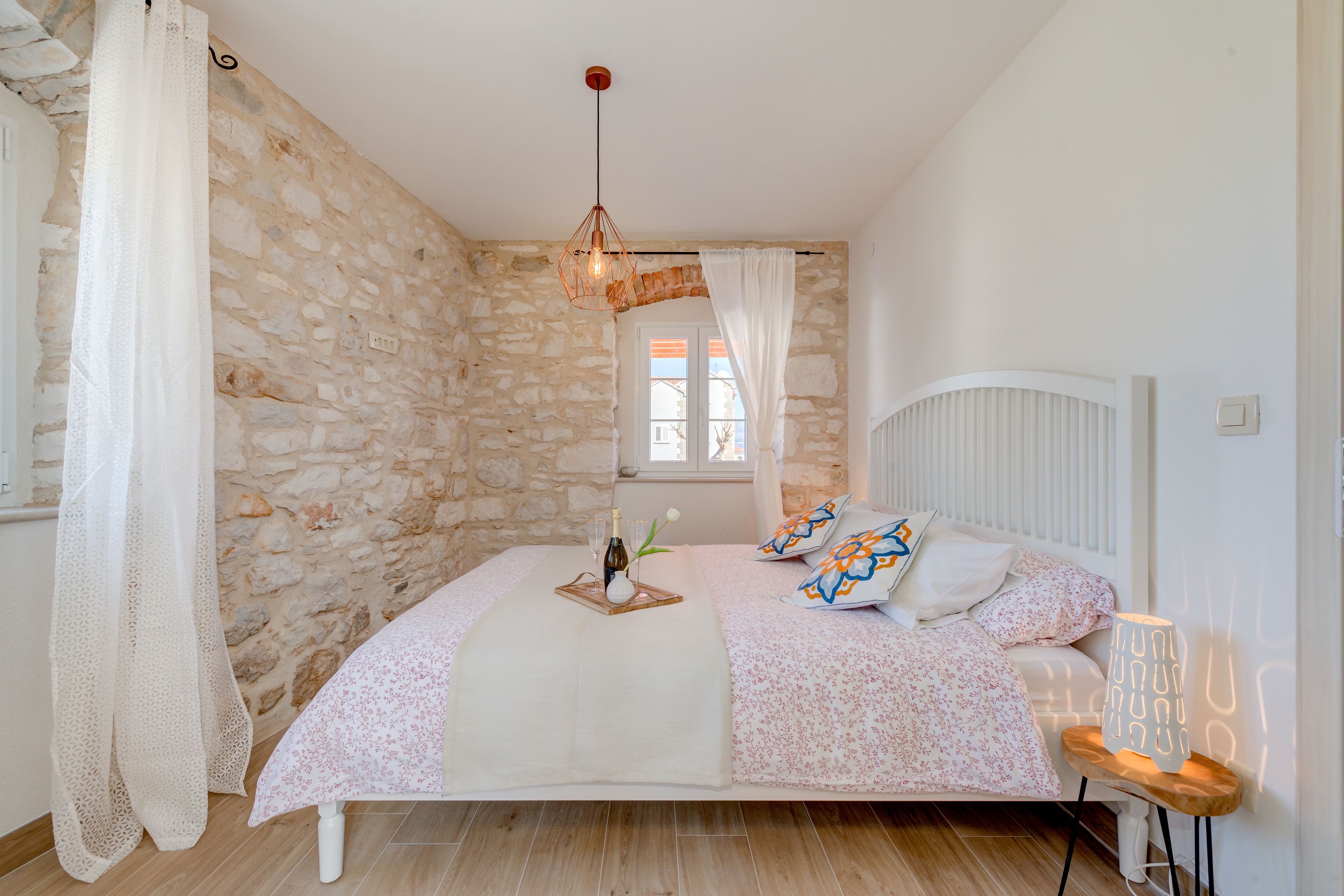 Une chambre avec un mur en pierre, un lit recouvert d’une literie à motifs floraux, une table de chevet avec une lampe et une fenêtre avec des rideaux.