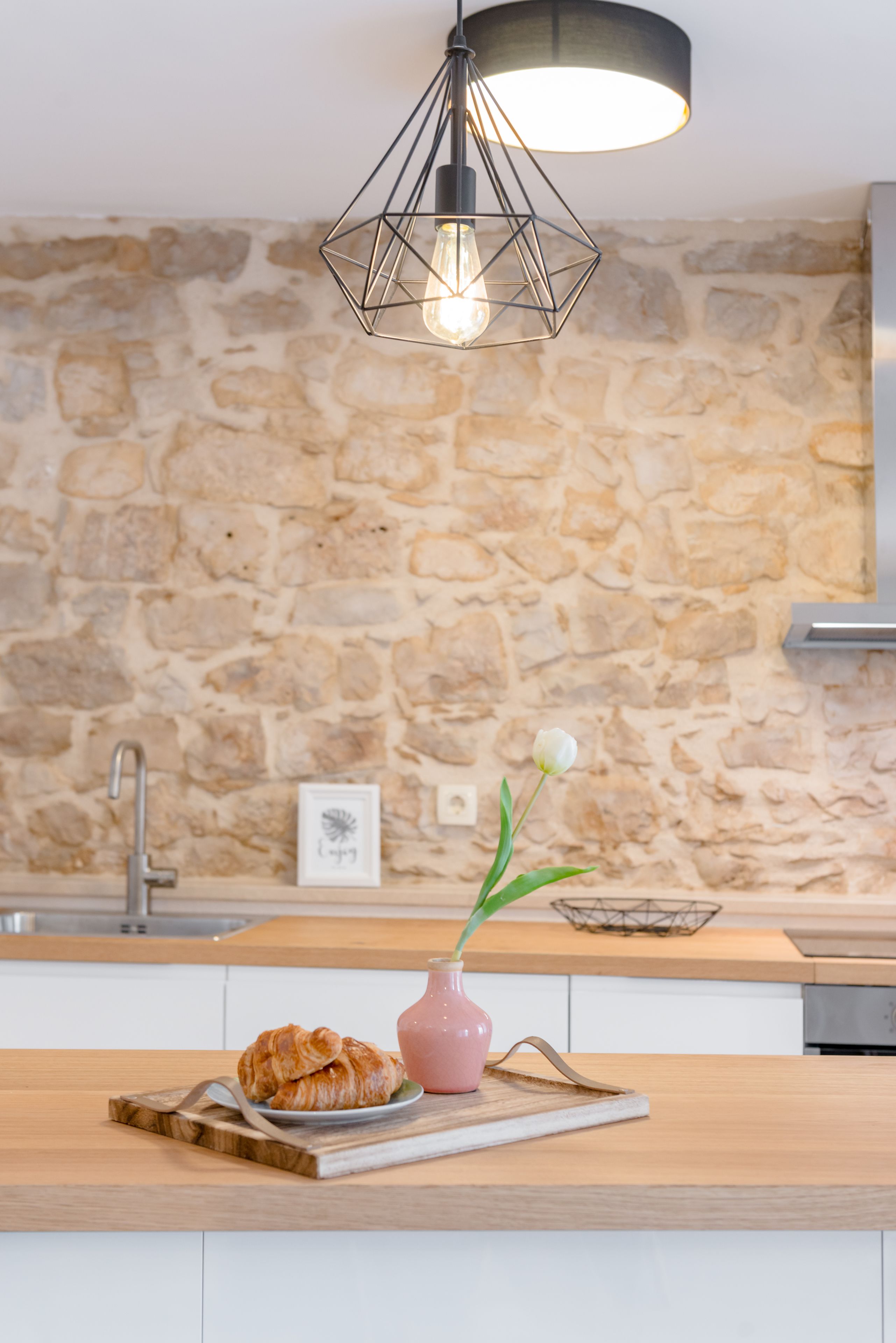 A kitchen with a stone wall, a wooden countertop, a pink vase with a white flower, and a plate with croissants.